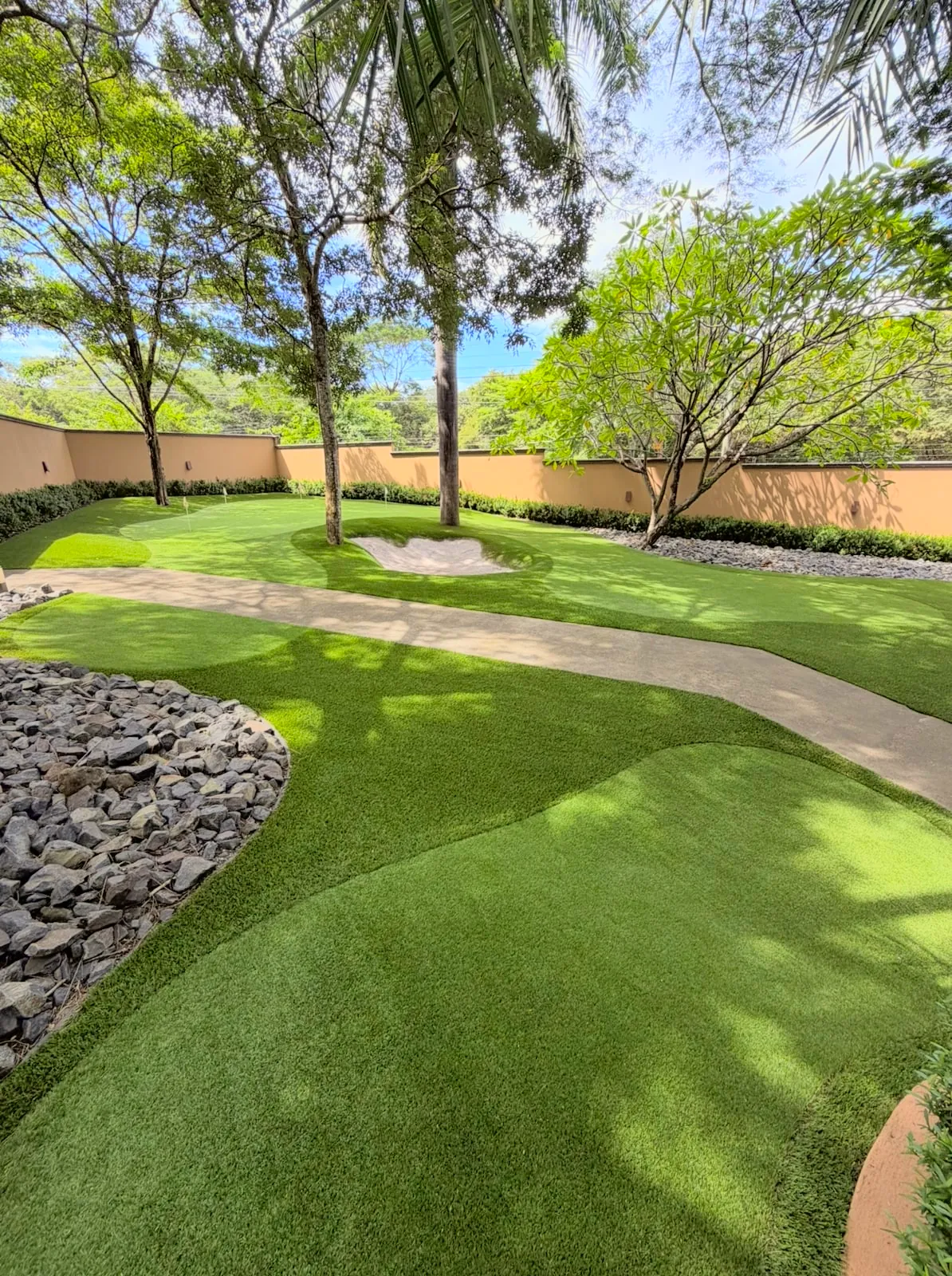 A shaded garden putting green with a sand bunker ringing a tree and a curving path