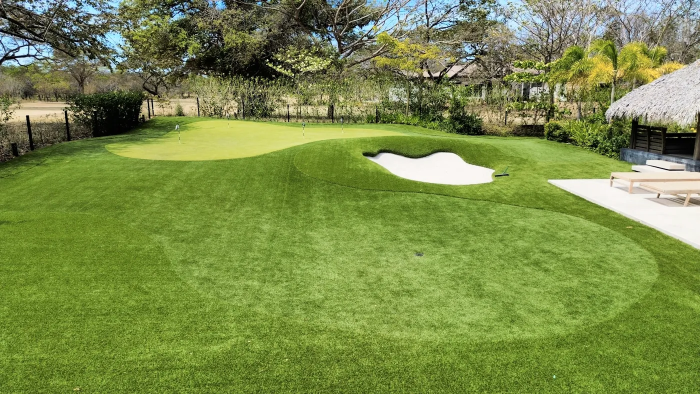 A putting green and sand bunker set into a wide lawn under shade trees