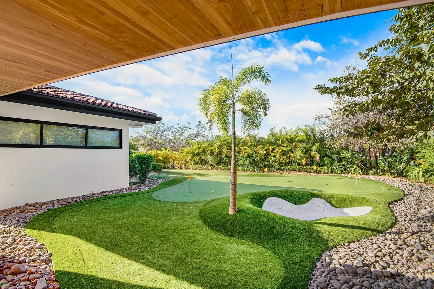 A putting green wrapped around a palm and a sand bunker, seen from a shaded terrace