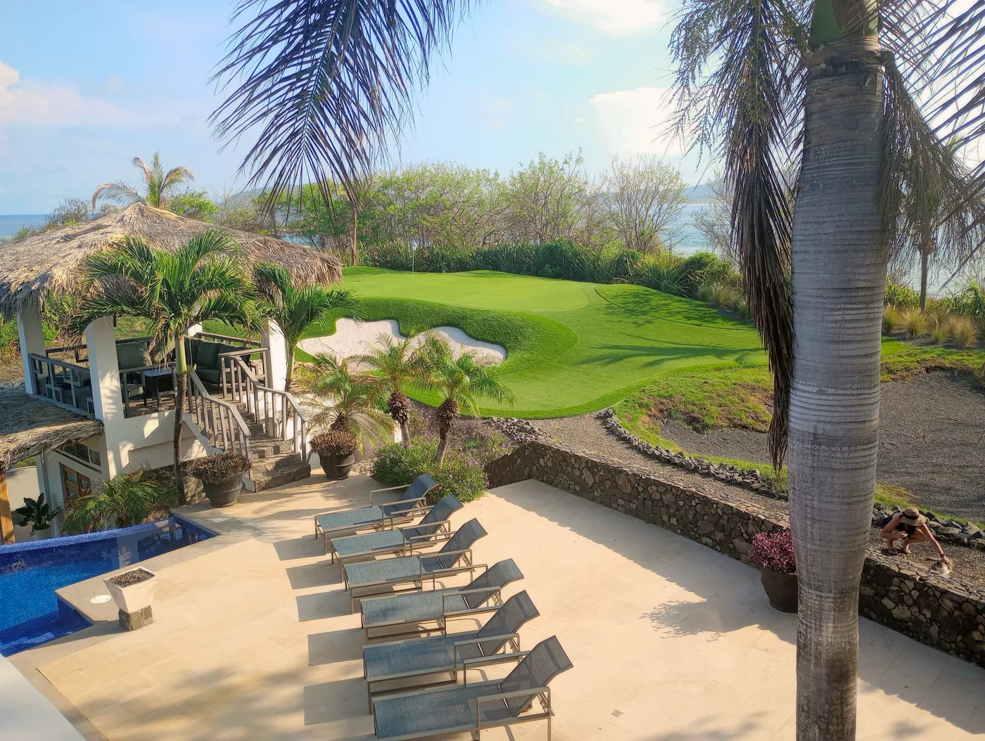 A pool deck lined with loungers below a hillside putting green and bunker