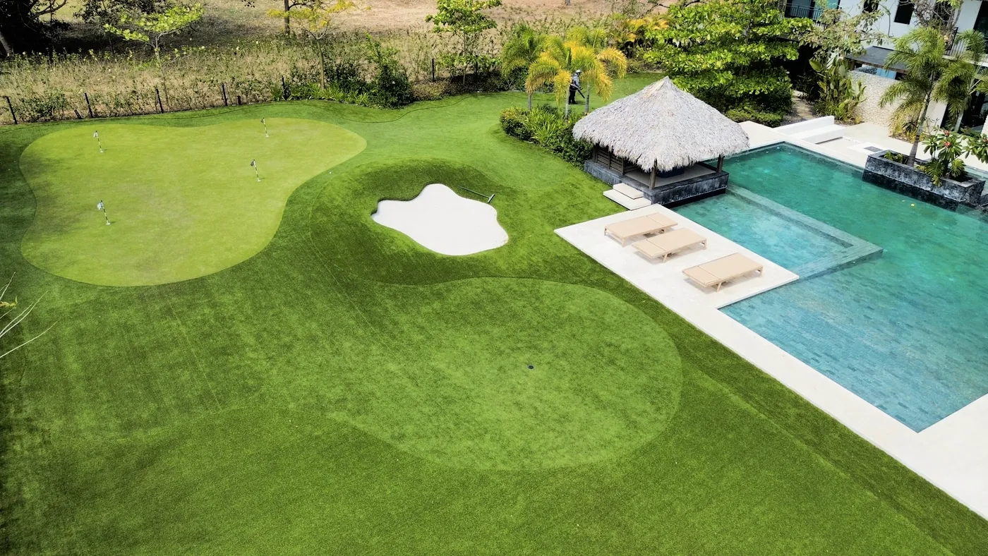Drone view of a pool deck with loungers and a palapa beside a putting green